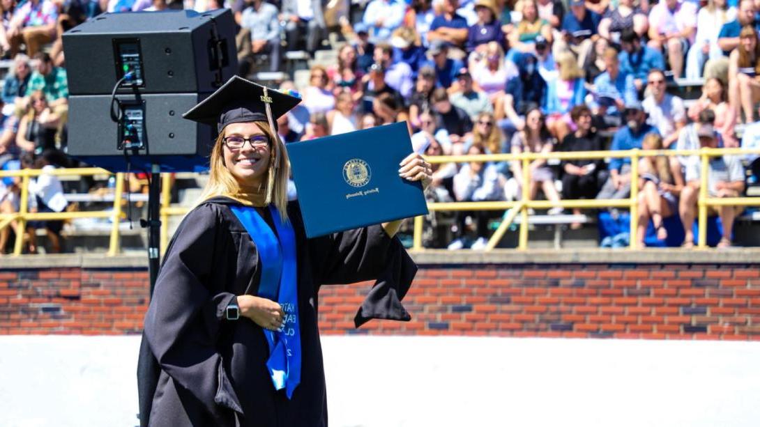 A student in a cap and gown holds up a diploma at Commencement